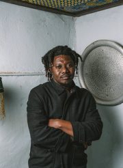 The author wearing a long-sleeved grey sweater, leaning against a wall with his arms folded.