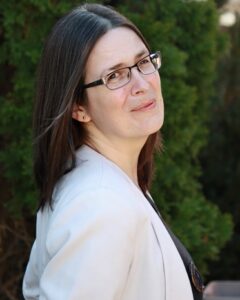 A white woman with shoulder-length straight dark brown hair and glasses, wearing a white blazer. She's looking at the camera over her right shoulder, in front of a leafy background.