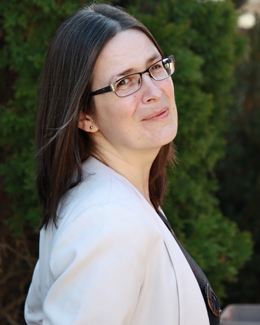 A white woman with shoulder-length straight dark brown hair and glasses, wearing a white blazer. She's looking at the camera over her right shoulder, in front of a leafy background.