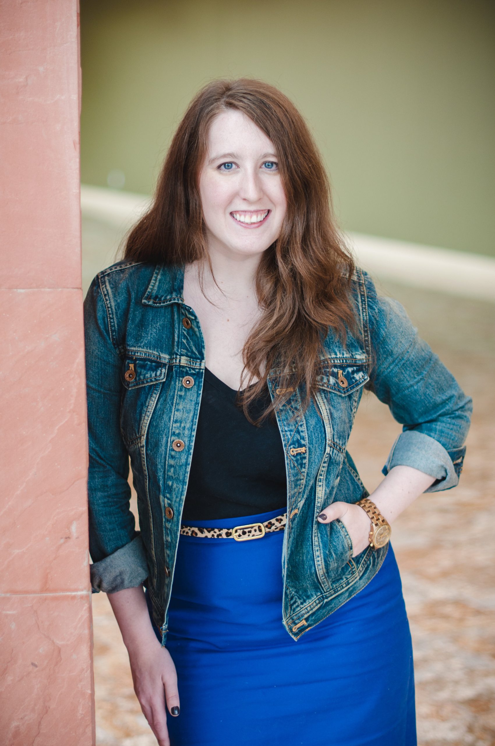 A photo of Melissa Frain. She is wearing a jean jacket over a black top and blue skirt, and leaning against a wall outdoors. She has one hand in her pocket.